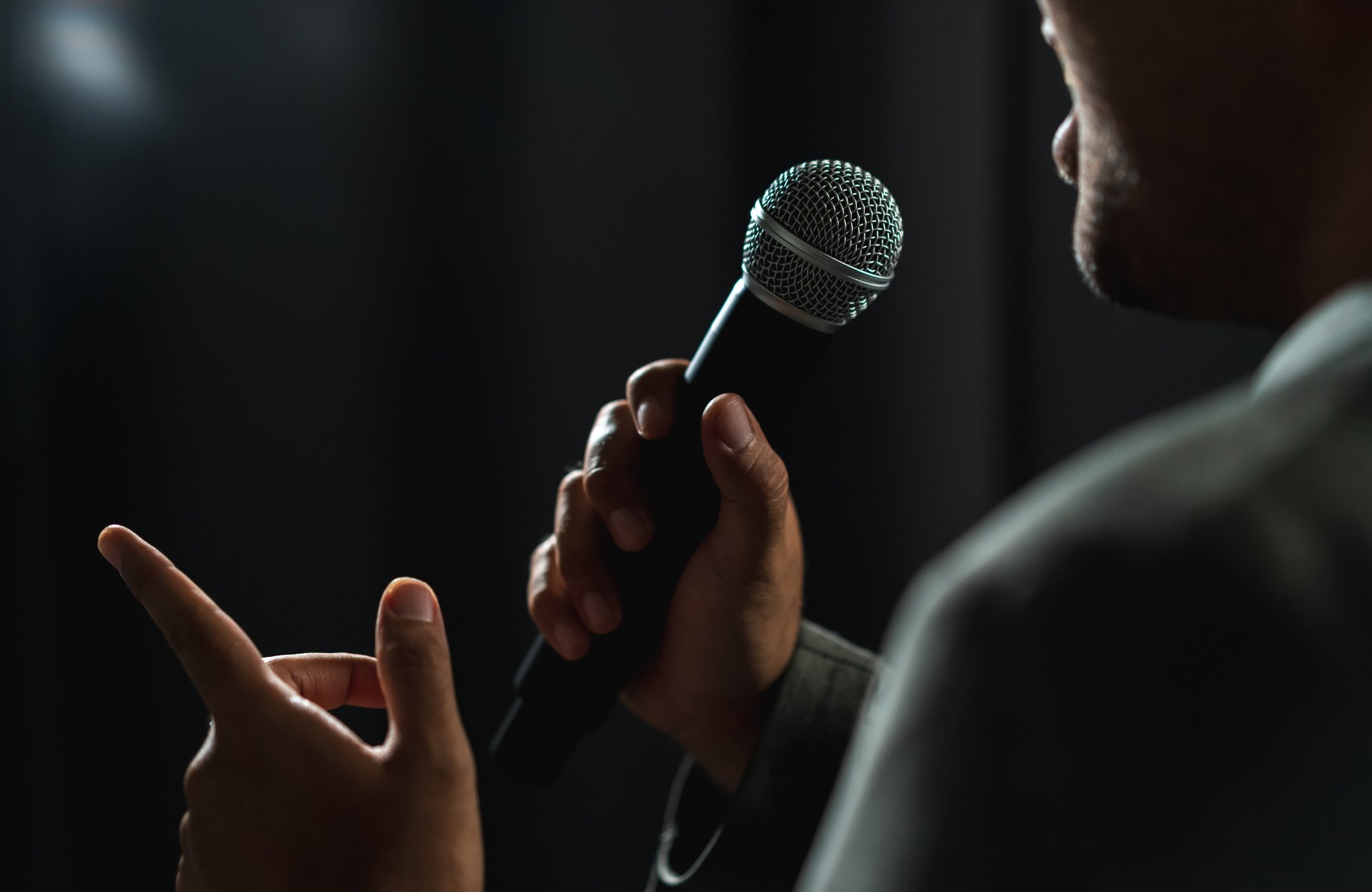 Confident successful speaker man talking on stage with spotlight strike through the darkness at corporate business event. Public speaker giving talk at conference hall. Stand up comedian.